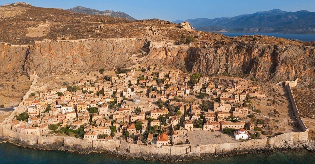 A coastal town with a fortified wall, dense buildings, and a mountainous backdrop. 3