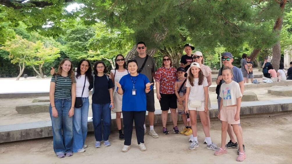 A group of people standing outdoors, possibly at a park or public space with trees and stone structures in the background. 1