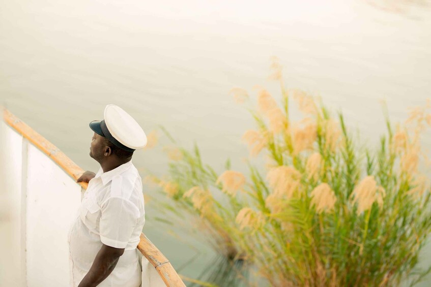 A person in a white uniform and hat standing by a railing, overlooking a body of water with tall grasses. 4