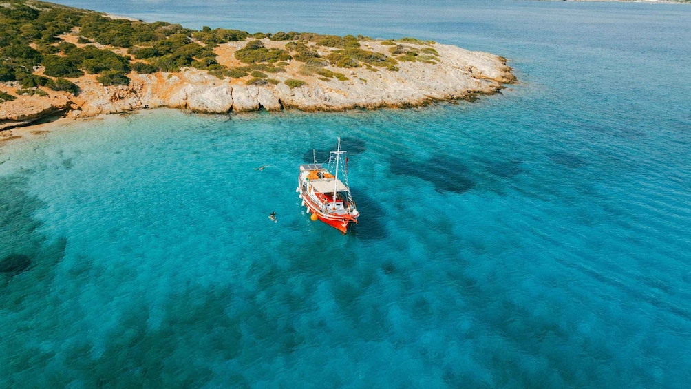 A red boat anchored in clear turquoise waters near a rocky coastline with sparse vegetation. 1
