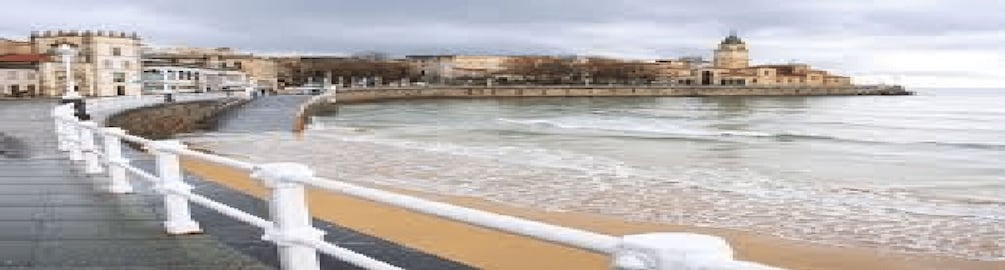 A coastal promenade with a white railing, a sandy beach, and historic buildings in the background. 4
