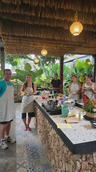 A group of people dining outdoors under a thatched roof, with a stone counter and various cooking utensils visible. 3