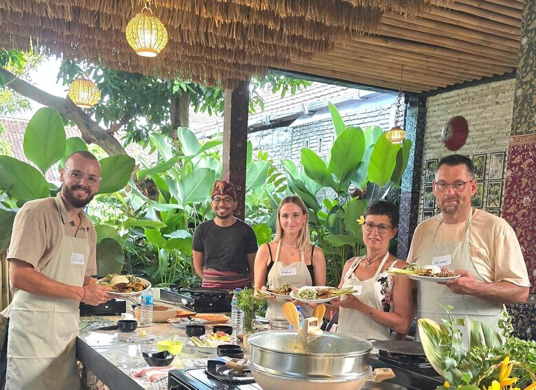 A group of people standing around a table with food, in an outdoor setting with plants and a thatched roof. 1