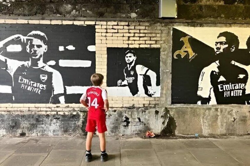 A child in a red sports jersey standing in front of a mural with black and white images of soccer players. 4
