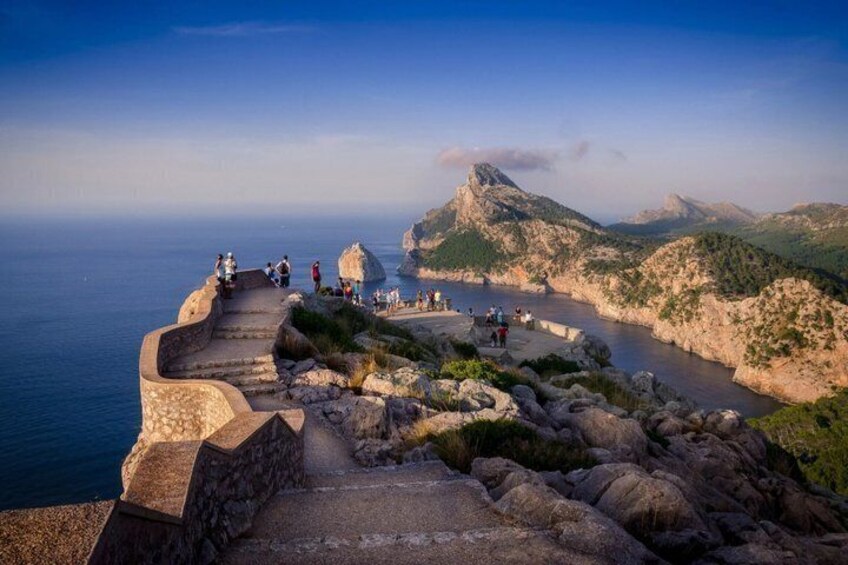A coastal path with people walking, a rocky shoreline, and a mountain in the background. 1