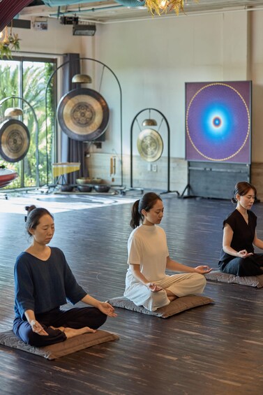 Three people meditating in a room with wooden floors, large windows, and decorative plates. 5