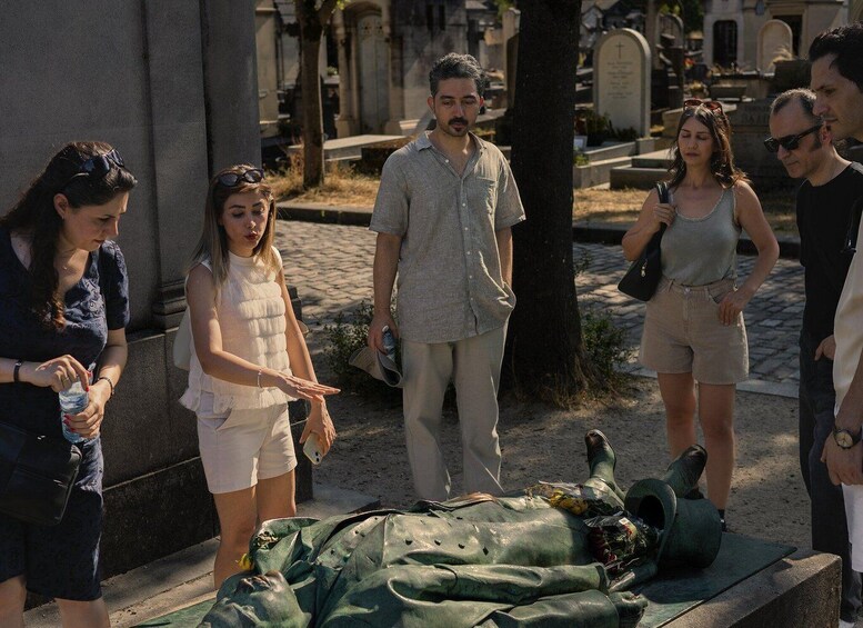 A group of people gathered around a green military uniform laid out on a stone surface in a cemetery with tombstones in the background. 2