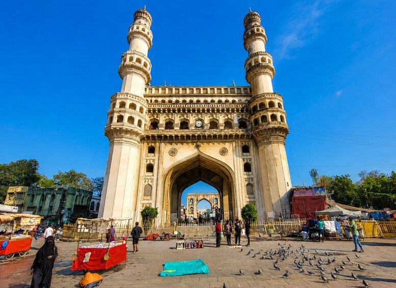 A historic archway with two towers, surrounded by a bustling market area with vendors and pigeons. 1