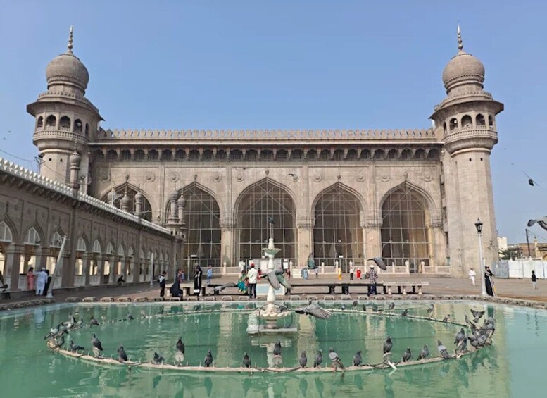 A historic building with large arched windows and two domed towers. In front, there is a fountain with birds and people around it. 2