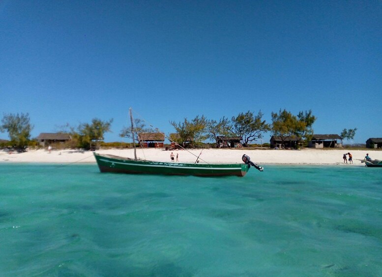 A green boat on clear turquoise water near a sandy beach with huts and sparse vegetation. 1