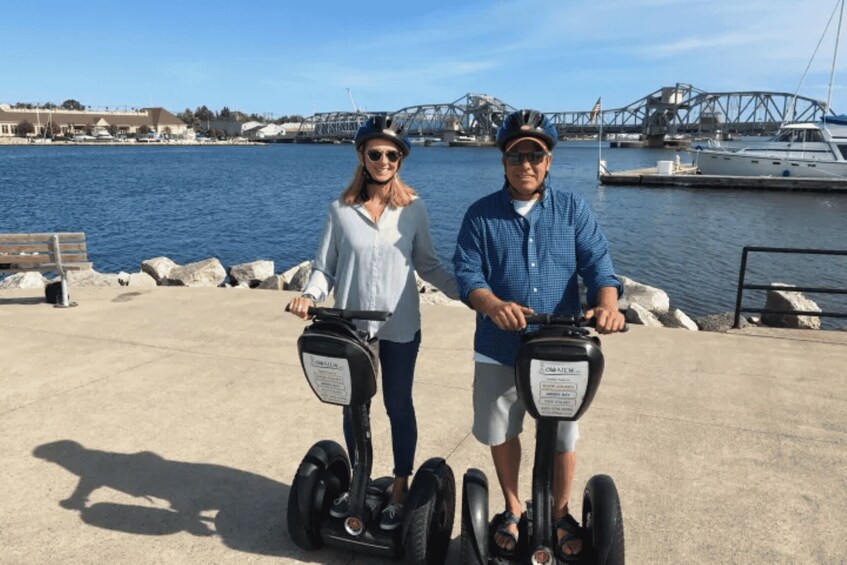 Two people on Segways near a waterfront with boats and a bridge in the background. 5
