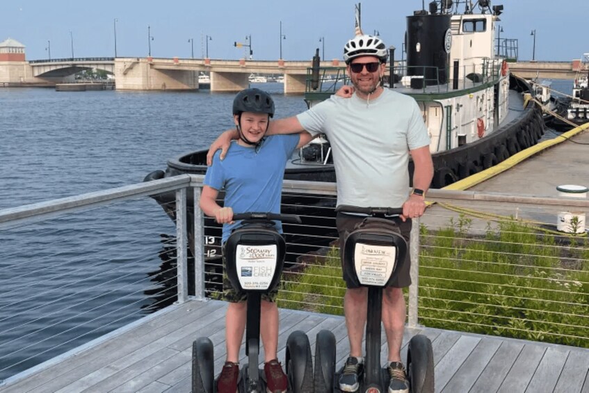 Two people on electric scooters by a waterfront with a bridge in the background. 3