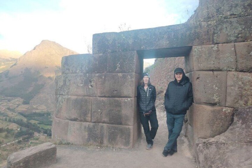 Two people standing in front of a stone structure with a mountainous landscape in the background. 2