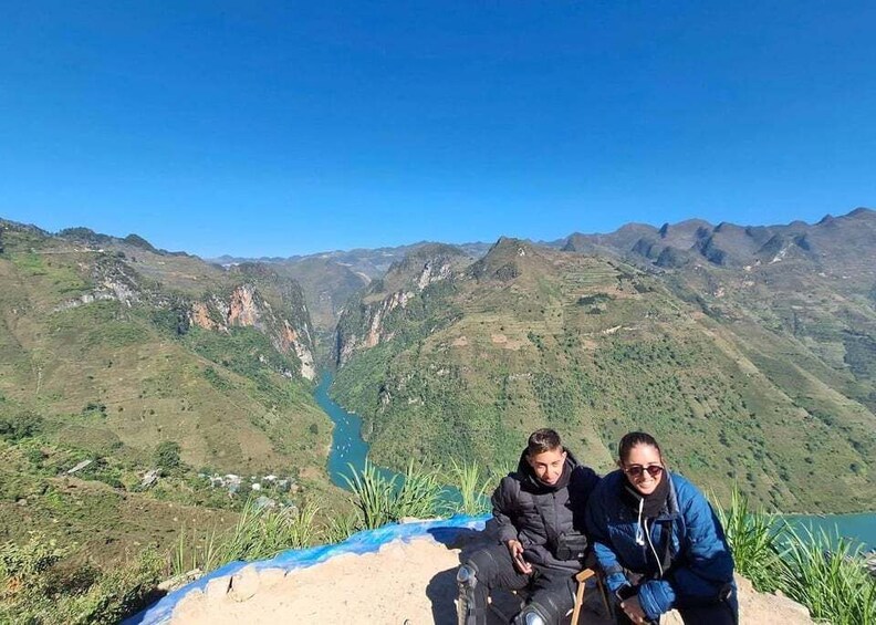 Two people sitting on a ledge overlooking a valley with a river and mountains in the background. 2