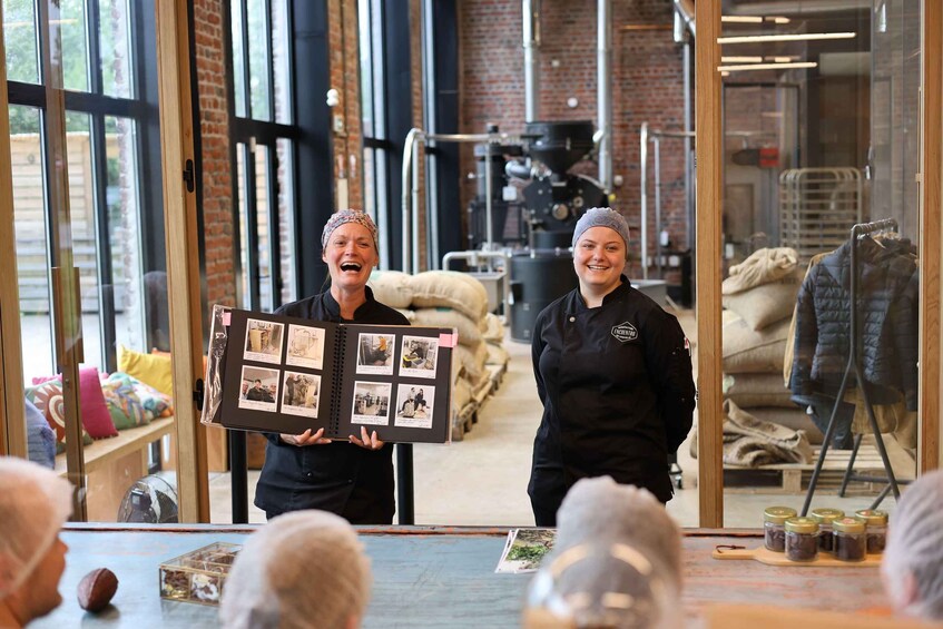 Two individuals holding a photo album in a rustic interior with brick walls and large windows. 3