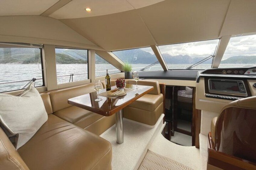 A boat interior with a dining area, cushioned seating, and a view of the water. 2