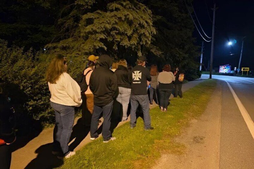 A group of people standing on a sidewalk at night, some wearing jackets. 2