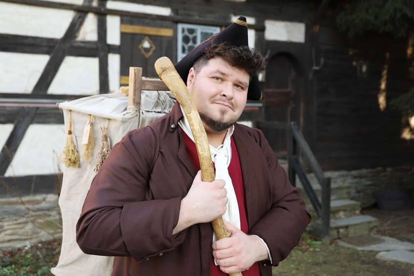 A person holding a wooden staff, wearing a brown jacket and a hat, standing in front of a building with a white and black facade. 1