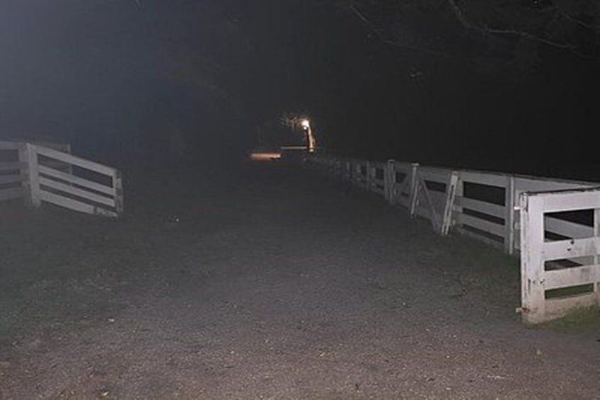 A dimly lit rural road with white fences on both sides and a light source in the distance. 3