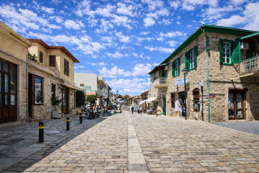 A cobblestone street lined with stone buildings, some with green shutters, under a partly cloudy sky. 1