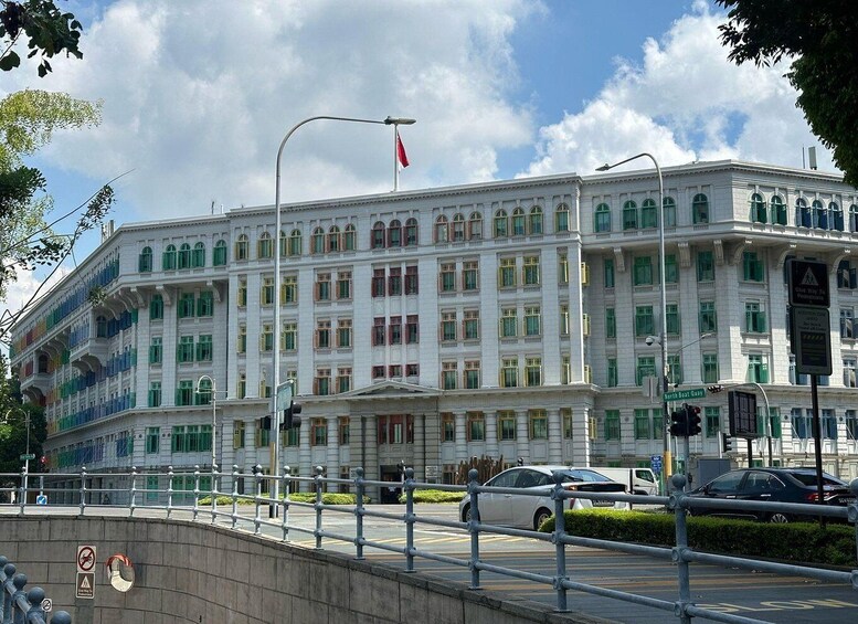 A large white building with green window frames and a red flag on top. There are cars parked in front and a pedestrian crossing. 5