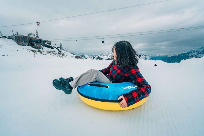 A person sitting on a snow tube on a snowy slope with ski lifts and mountains in the background. 3