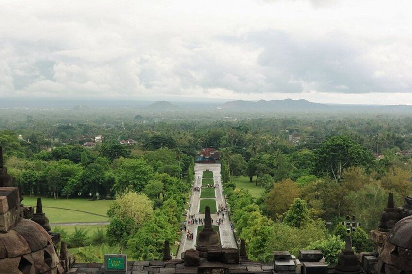 A view of a temple complex with a central pathway leading to a raised platform, surrounded by lush greenery and distant hills. 4