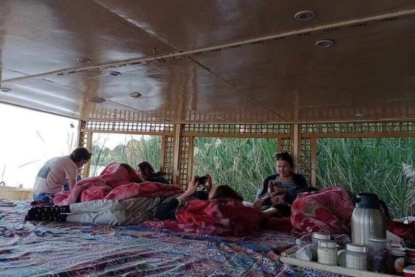 A group of people sitting on a patterned carpet under a wooden structure with a lattice design, surrounded by tall grass. 2