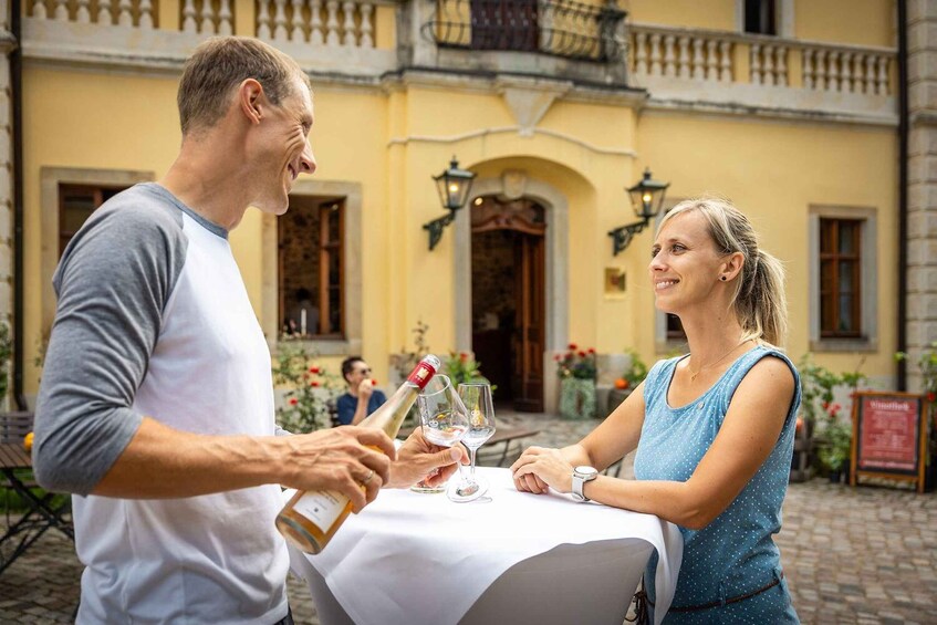 A man and a woman toasting with wine glasses at an outdoor table with a yellow building in the background. 3