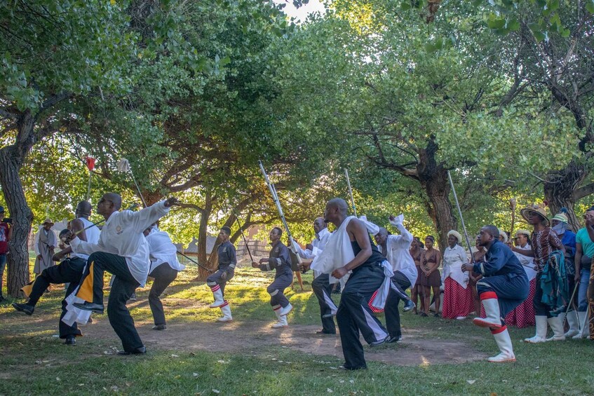 A group of people performing a traditional dance in an outdoor setting with trees and grass. 5