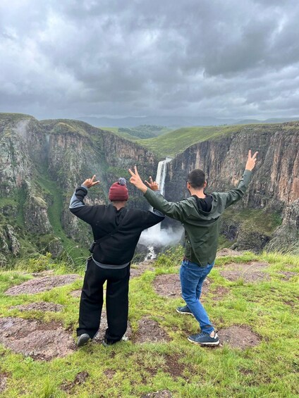Two people standing on a grassy hill overlooking a waterfall. 1