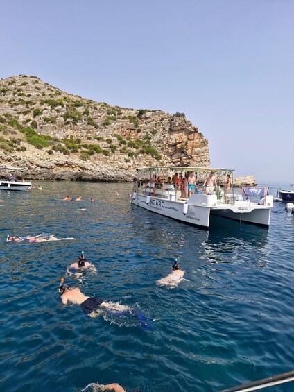 A boat named 'INGARO' with people swimming and snorkeling in the water, a rocky cliff in the background, and a clear sky. 4