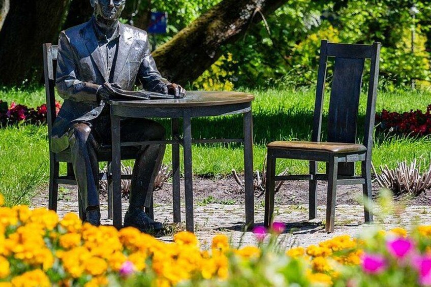 A statue of a seated man in a suit, a wooden chair, and a table in a park with flowers. 5