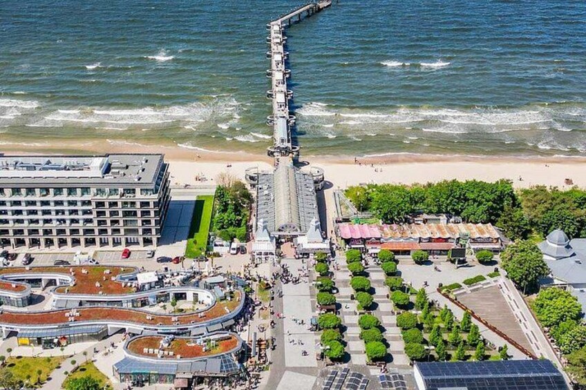 A coastal cityscape with a pier extending into the sea, a modern building complex, and a landscaped area with trees. 1