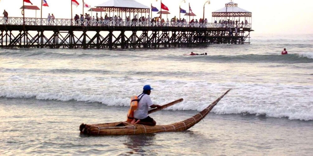 A person paddling a long wooden boat on a beach with waves, a pier with flags, and a crowd in the background. 1