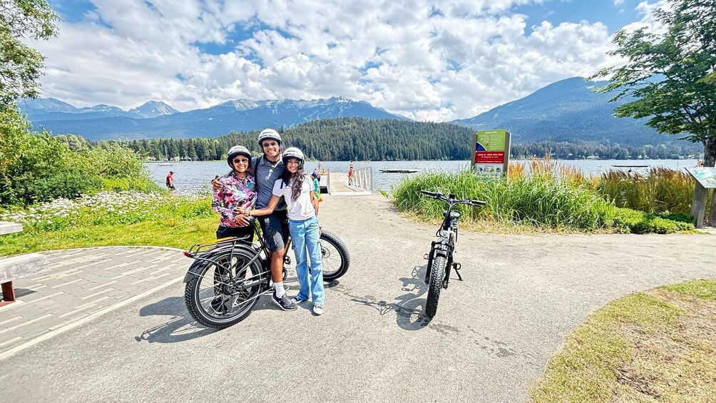 Three people posing with bicycles in a scenic outdoor area with mountains and a lake in the background. 4