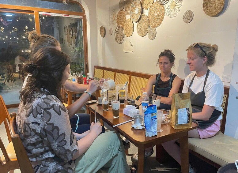 Three people sitting at a wooden table with various items like cups, a bag, and a bottle. 3