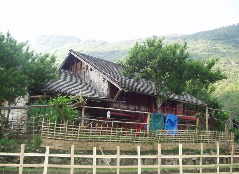 A wooden house with a tiled roof, surrounded by a bamboo fence, set against a backdrop of green hills. 1