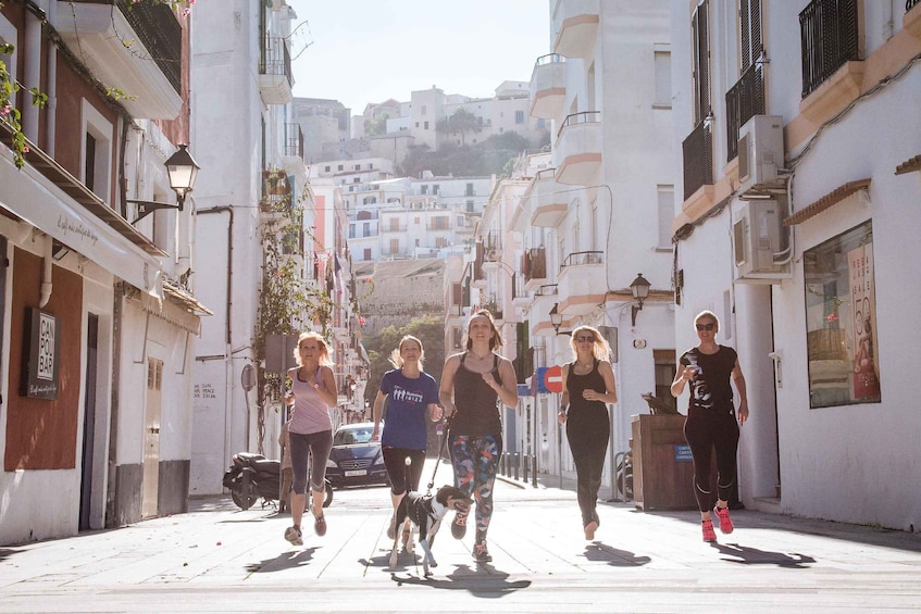 A group of people jogging down a narrow street with buildings on either side and a hilly landscape in the background. 4