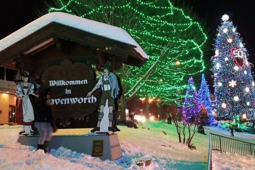 A snow-covered welcome sign with illuminated green lights, a Christmas tree, and a snowman figure. 5