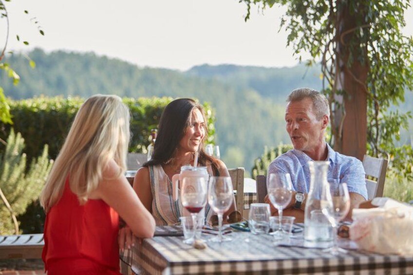 A group of people sitting at an outdoor table with wine glasses, surrounded by greenery and hills in the background. 3