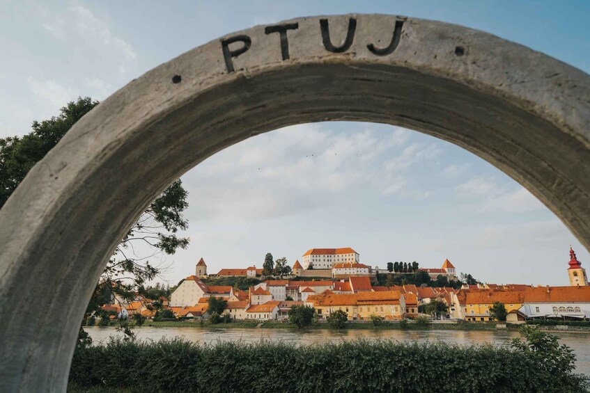 A stone arch with the inscription 'PTUJ' over a river, with buildings and a tower in the background. 4