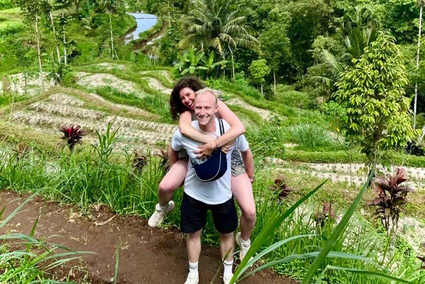 A person carrying another on their back in a lush, green landscape with terraced fields and a body of water in the background. 1