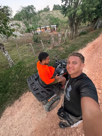 Two people on a dirt road with a vehicle, surrounded by greenery and a hill with buildings in the background. 2