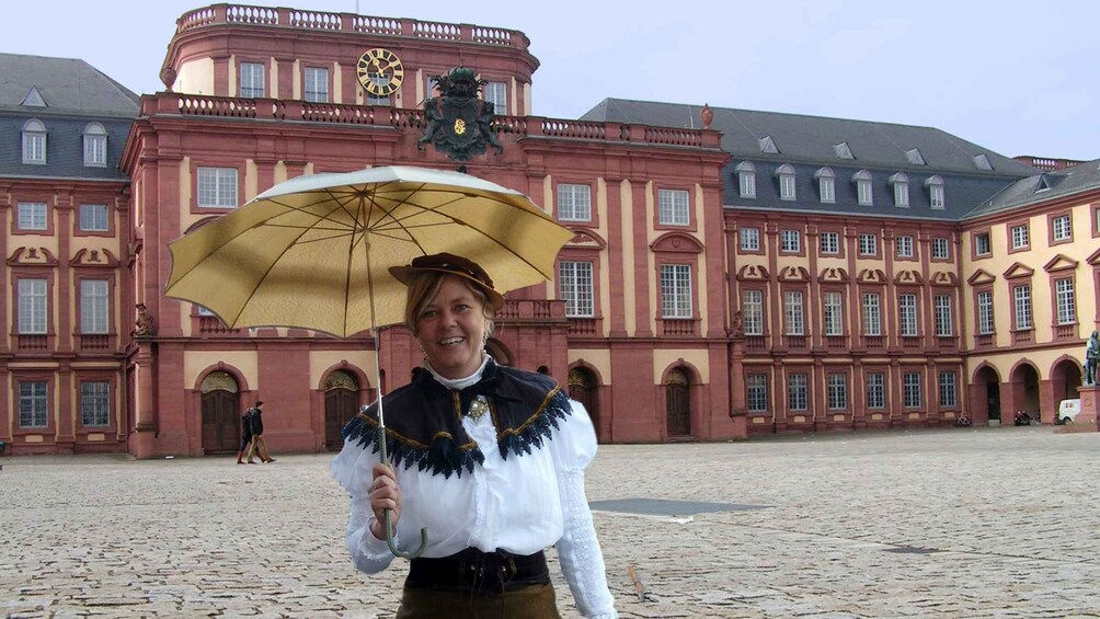 A person in traditional attire holding an umbrella in front of a historic building with a clock and a coat of arms. 1