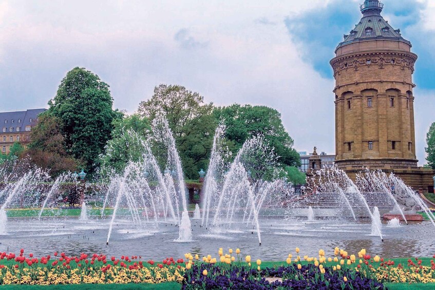 A fountain with water jets, surrounded by colorful tulips and flowers, with a historic tower in the background. 5