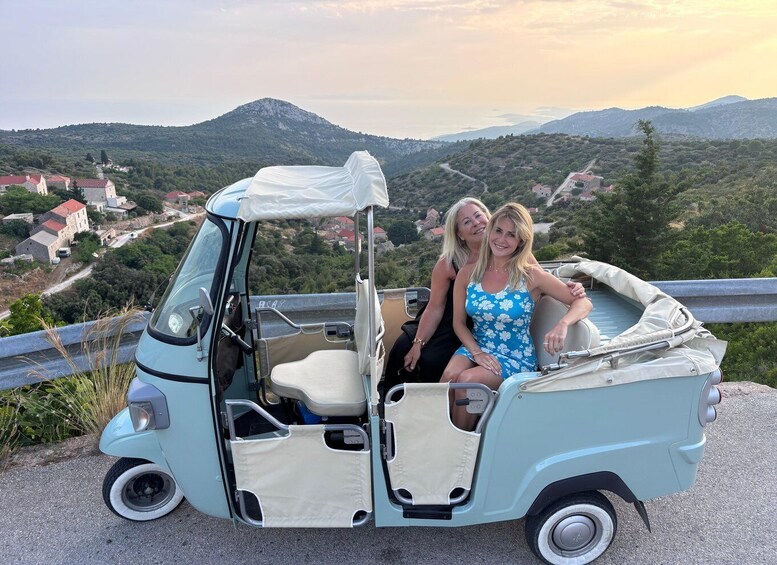 A light blue golf cart with two people seated, parked on a road with a scenic view of a mountainous landscape and houses in the background. 4
