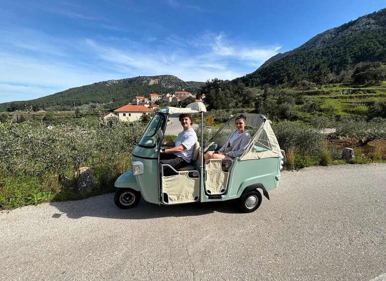 A vintage green car parked on a gravel road with two people inside, surrounded by a rural landscape with houses and hills in the background. 1