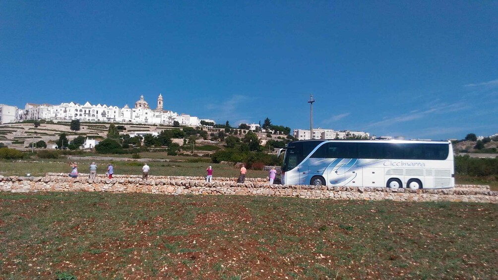 A white bus parked in a field with a stone wall, people walking, and a white building with a tower in the background. 2