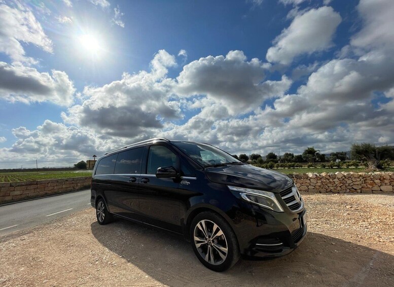 A black minivan parked on a dirt road with a stone wall and a field in the background under a partly cloudy sky. 3